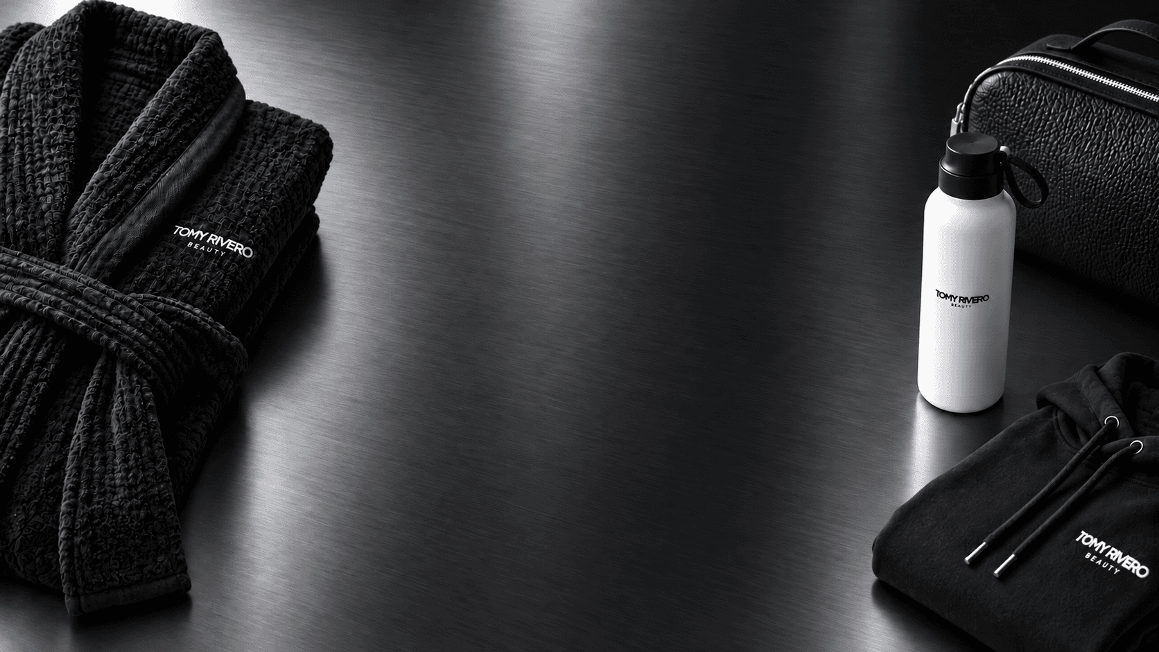 Black spa robe, textured travel case, white branded water bottle, and black hoodie arranged around an open charcoal and cool silver metallic surface