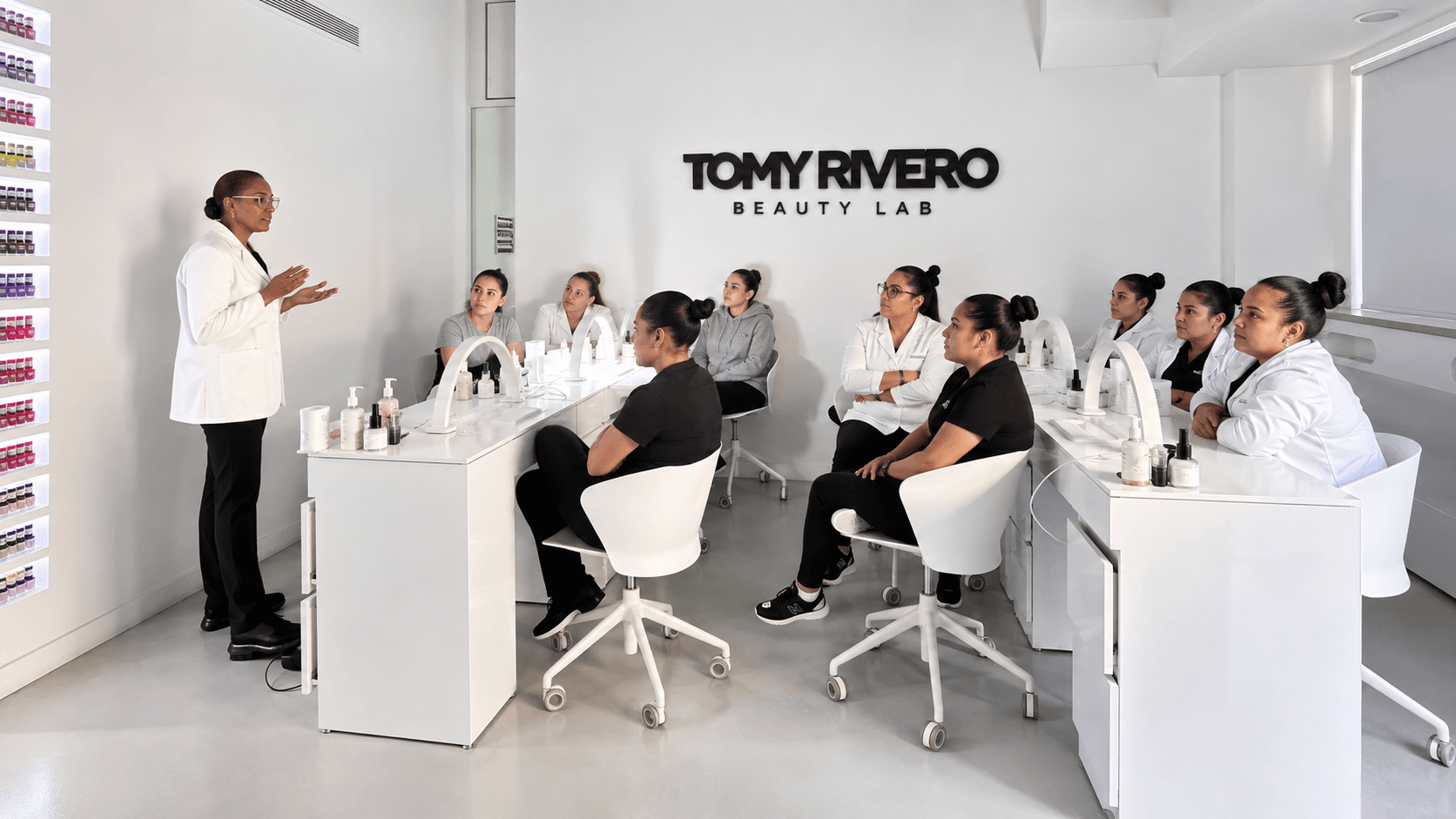 A Beauty Lab instructor leads a nail training class beneath the Tomy Rivero Beauty Lab wall logo